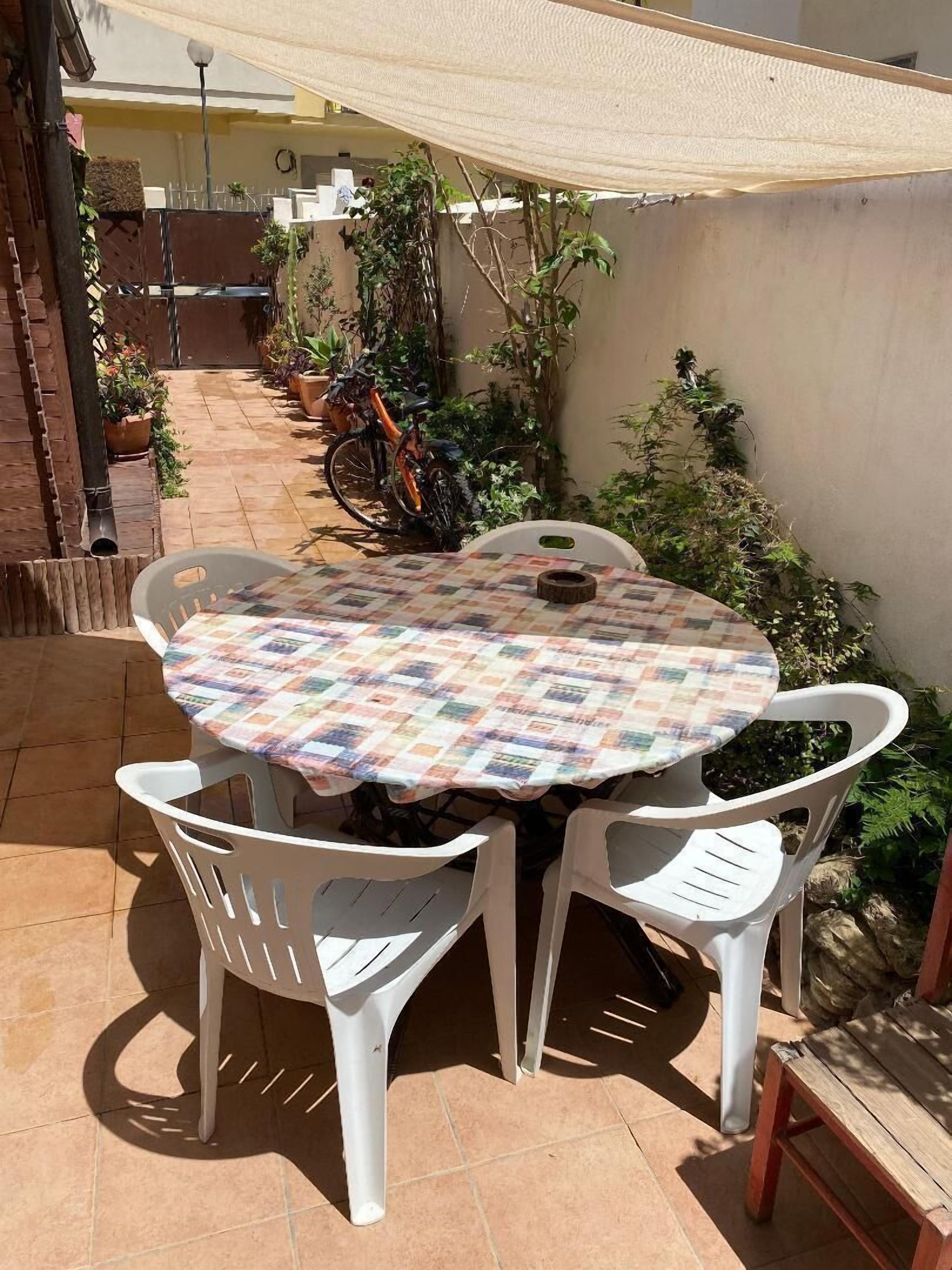 En udendørs patio med et rundt bord og hvide plastikstole, omgivet af krukker med planter og en cykel.