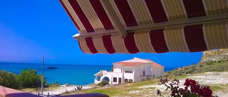 A coastal view with a building featuring a red-tiled roof and a white facade, under a striped awning.