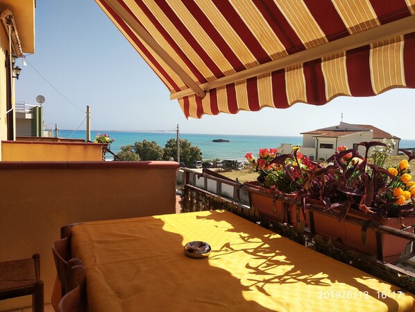 Terraço com mesa e cadeiras amarelas, com vista para a costa, onde se vê um edifício e um barco ao longe.