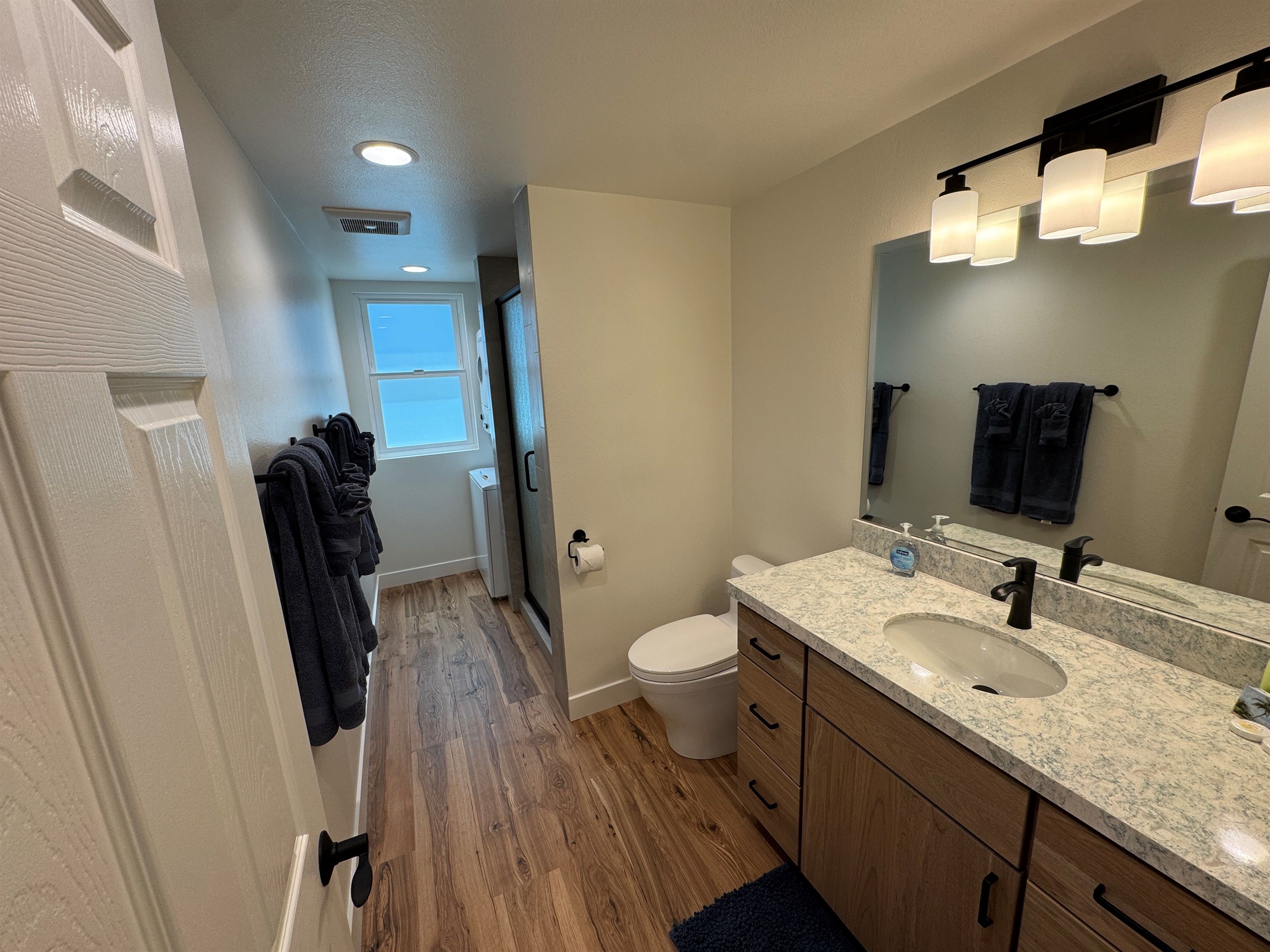Une salle de bain avec un plancher en bois, des toilettes blanches, un lavabo avec un comptoir en marbre et un miroir surmonté de luminaires.