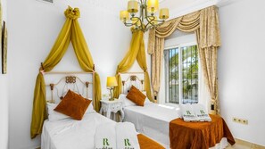 Two single beds with white linens and decorative headboards, a chandelier, and a large window with a view.