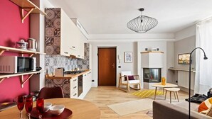 Een moderne keuken met een magnetron, fornuis en planken. Een eettafel met rode wijnglazen en kommen is zichtbaar. Een houten deur leidt naar een zithoek met een bank en een lamp.