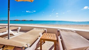Eine Strandlounge mit Liegestühlen und einem kleinen Tisch, mit Blick auf das klare, blaue Meer und einen fernen Steg.
