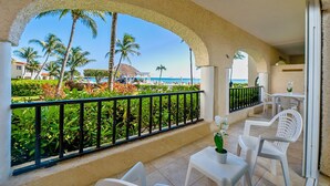 Ein Balkon mit weißen Stühlen, einem Tisch mit Topfpflanze und Blick auf einen Strand mit Palmen und einem Pool.