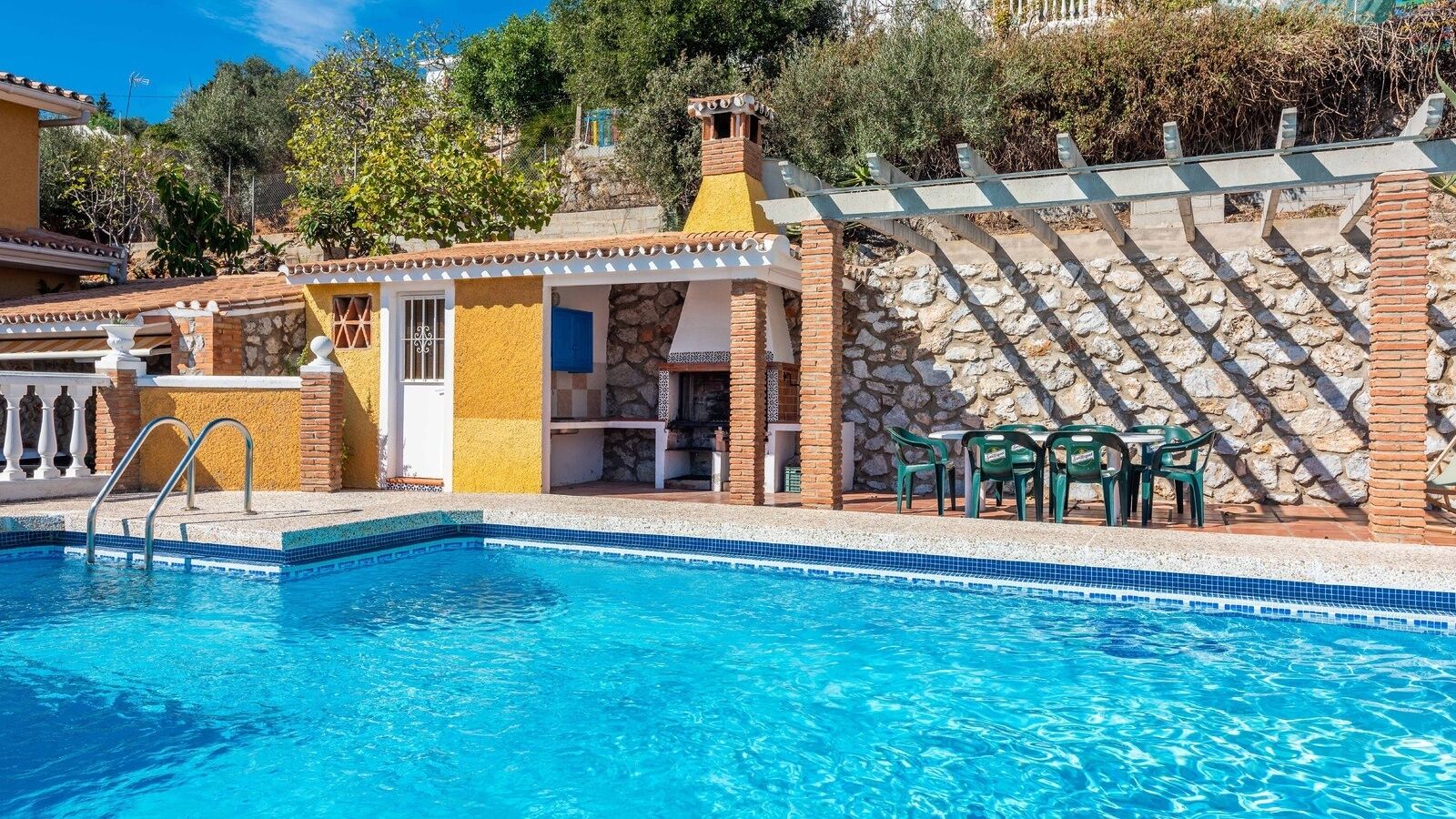 A swimming pool with a stone wall and a covered seating area with chairs.
