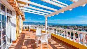 Ein Balkon mit weißen Korbstühlen und einem Tisch, mit Blick auf eine Stadtlandschaft unter blauem Himmel.