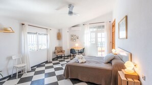 Ein Hotelzimmer mit Schachbrettmuster auf dem Boden, einem Bett mit Kissen, einem Schreibtisch mit Stuhl, einem zusätzlichen Stuhl, einem Deckenventilator und einem Fenster mit Vorhängen.