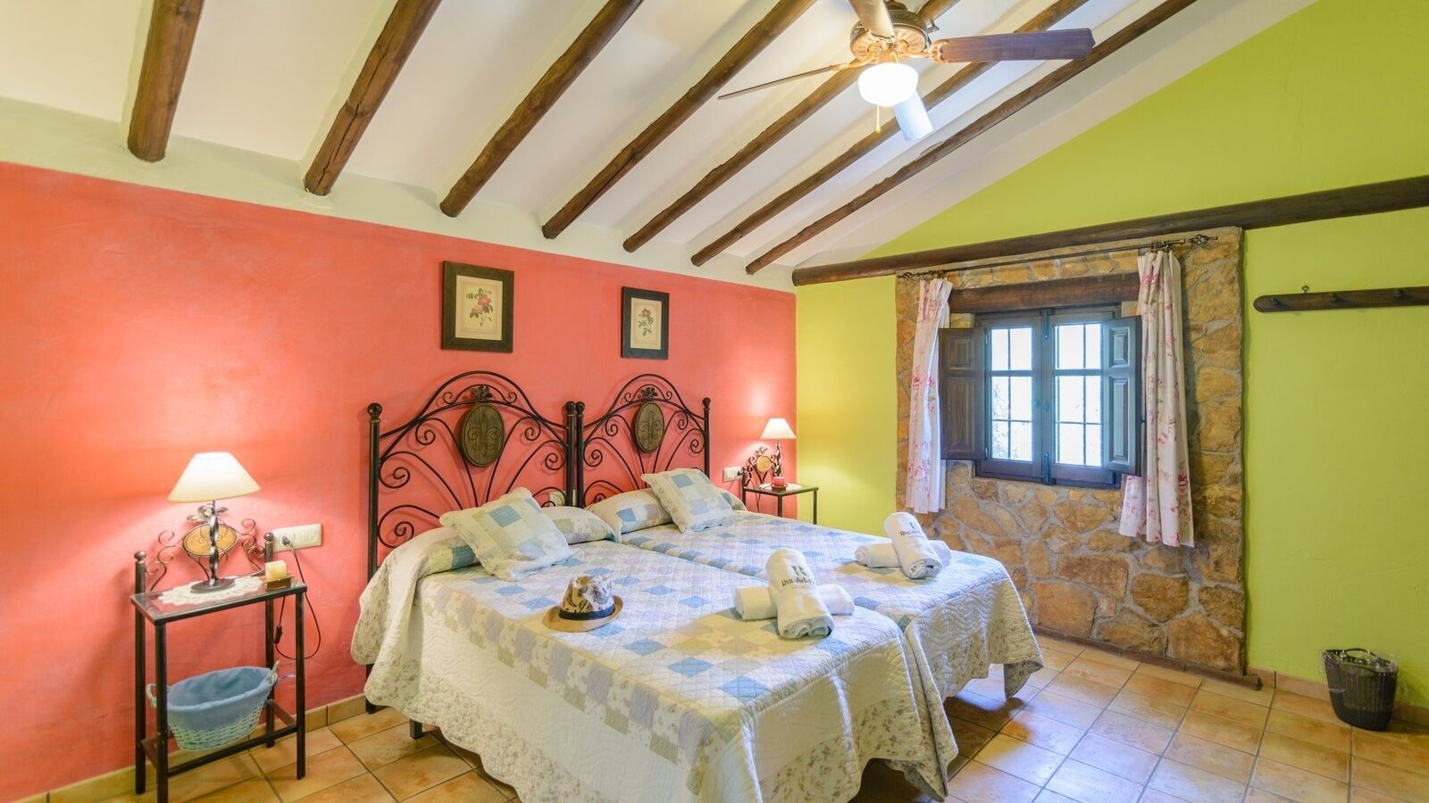 A bedroom with a bed, two bedside tables, a window with curtains, a stone wall, and a ceiling fan.
