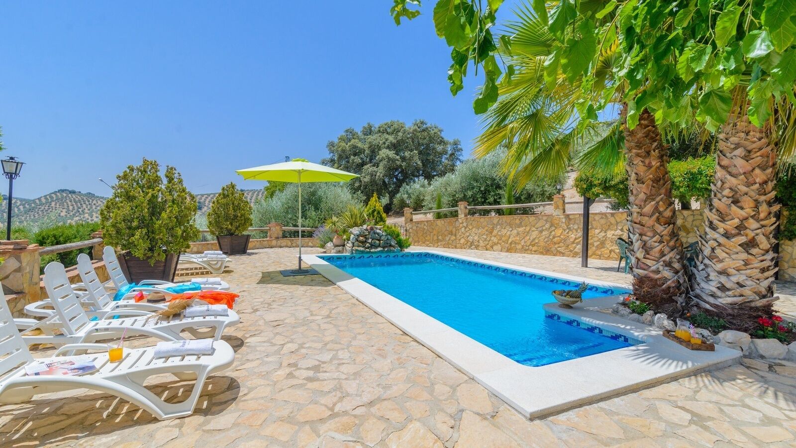 A swimming pool with lounge chairs and a yellow umbrella, surrounded by palm trees and stone walls.