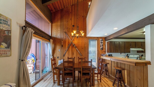 A wooden dining area with a long table and chairs, a chandelier, and a kitchen visible through an open door. - 1+Loft on Ski Back Trail at Canyon Lodge w/ Garage! (Mammoth Lakes)