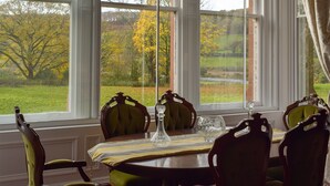 Uma sala de jantar com uma mesa posta para quatro pessoas, janelas amplas e vista para uma paisagem verde com árvores.