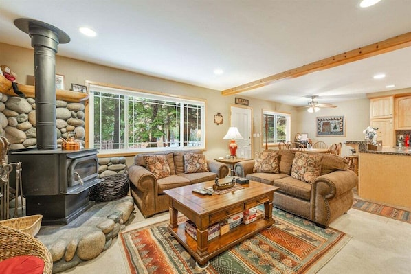 A cozy living room with a stone fireplace, two sofas, a coffee table, and a kitchen area visible through an open door.
