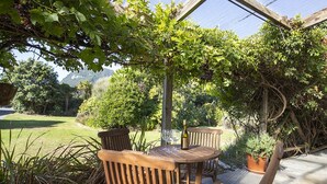 A wooden table and chairs under a pergola with grapevines, surrounded by lush greenery and a view of a mountain. - Cliffscapes Punakaiki (Punakaiki)