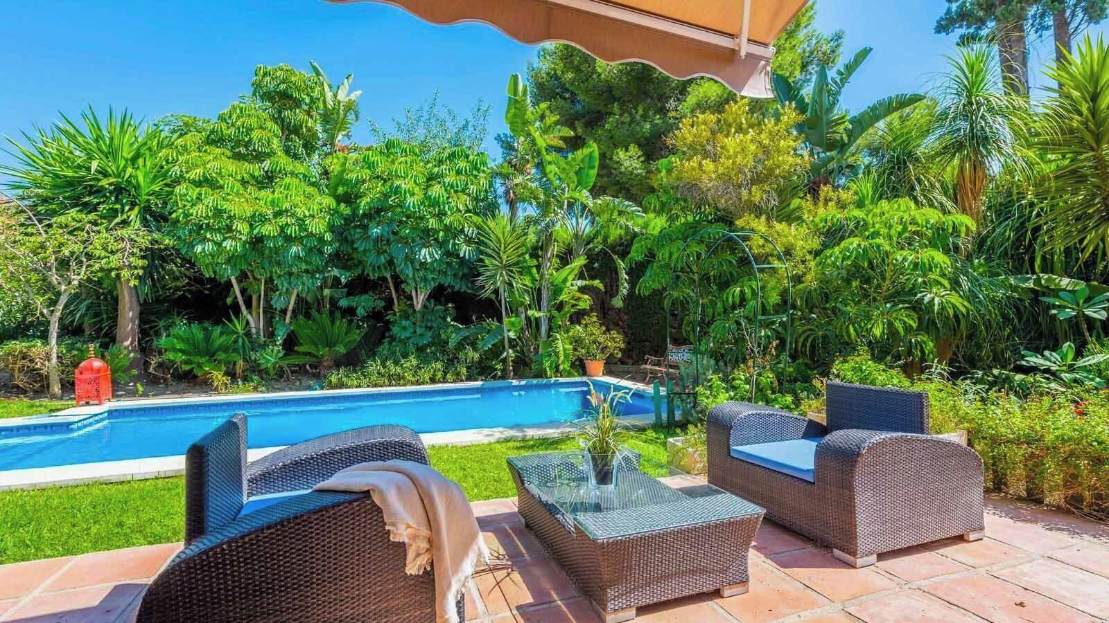 A poolside area with wicker furniture and a clear view of a swimming pool surrounded by lush greenery.