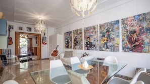 A dining area with a glass table, white chairs, and a white sofa. There are guitars and a chandelier.