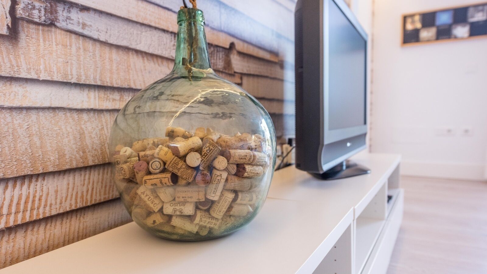 A glass bottle filled with wine corks on a white surface with a television in the background.