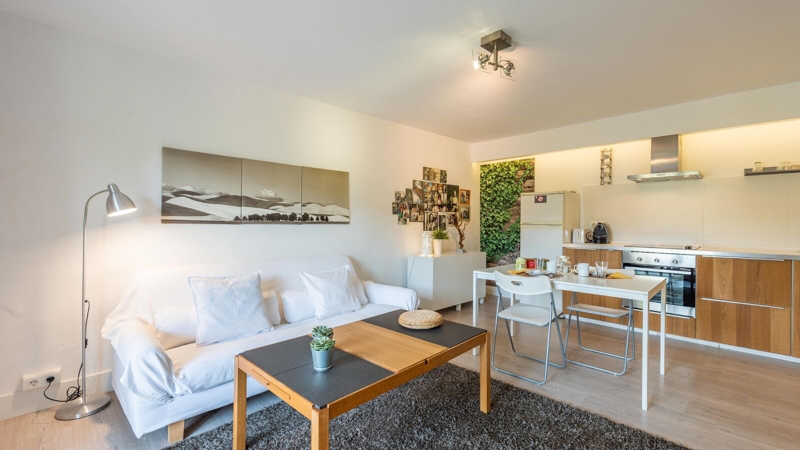 Un salon moderne avec un canapé, une table basse et un coin repas avec des chaises. On aperçoit une cuisine à l’arrière-plan, avec une cuisinière et des placards.