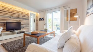 A modern living room with a flat-screen TV on a white stand, a wooden coffee table, a white sofa, and a large window with curtains. - Casa Ami Guaro by Ruralidays (Málaga)