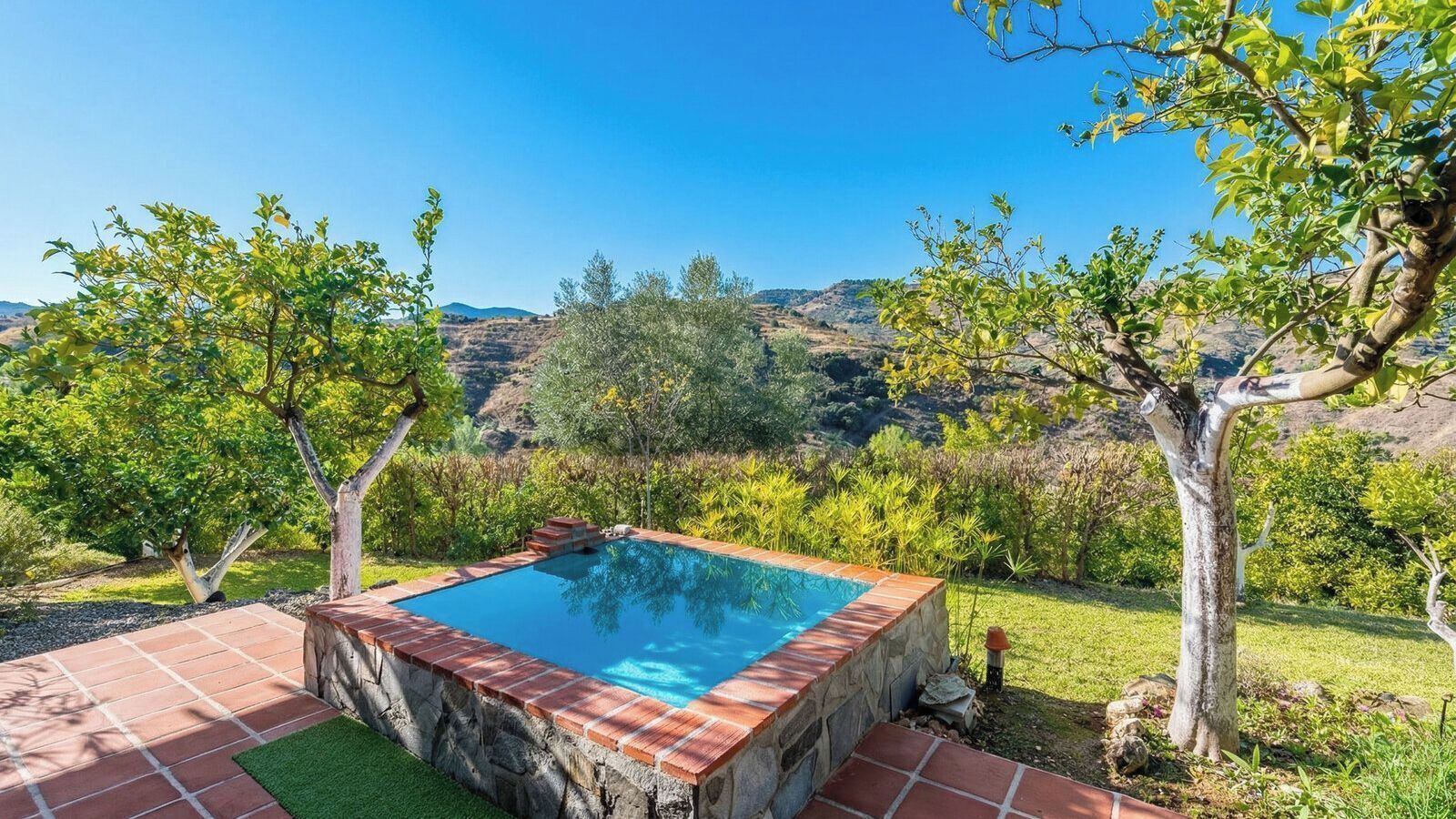 A rectangular pool with a stone border, surrounded by a grassy area and trees, with a hilly landscape in the background.