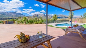 Eine Terrasse mit Holztisch, Obst und Getränken, ein Pool und Blick auf die Berge.