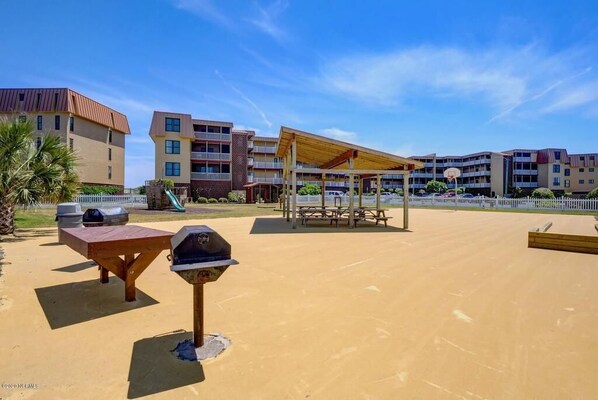 A sandy outdoor area with barbecue grills, picnic tables, and a covered seating area. - Oceanfront End Unit 2BR Condo - Linens Included (North Topsail Beach)