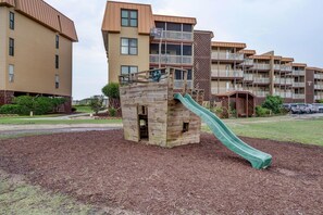 A playground with a wooden slide and climbing structure, surrounded by a landscaped area with mulch.