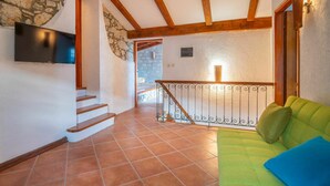 Amplia sala de estar con sofá verde, televisor montado en la pared, pared de piedra, vigas de madera y una escalera que conduce a un nivel superior.