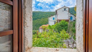 Un edificio rústico de piedra con fachada blanca y contraventanas azules, rodeado de exuberante follaje y un telón de montañas.