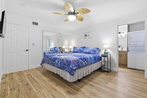 Ein Schlafzimmer mit Bett, Deckenventilator, Spiegel, einer Wand mit einem Motivationsspruch und einem Waschbereich.