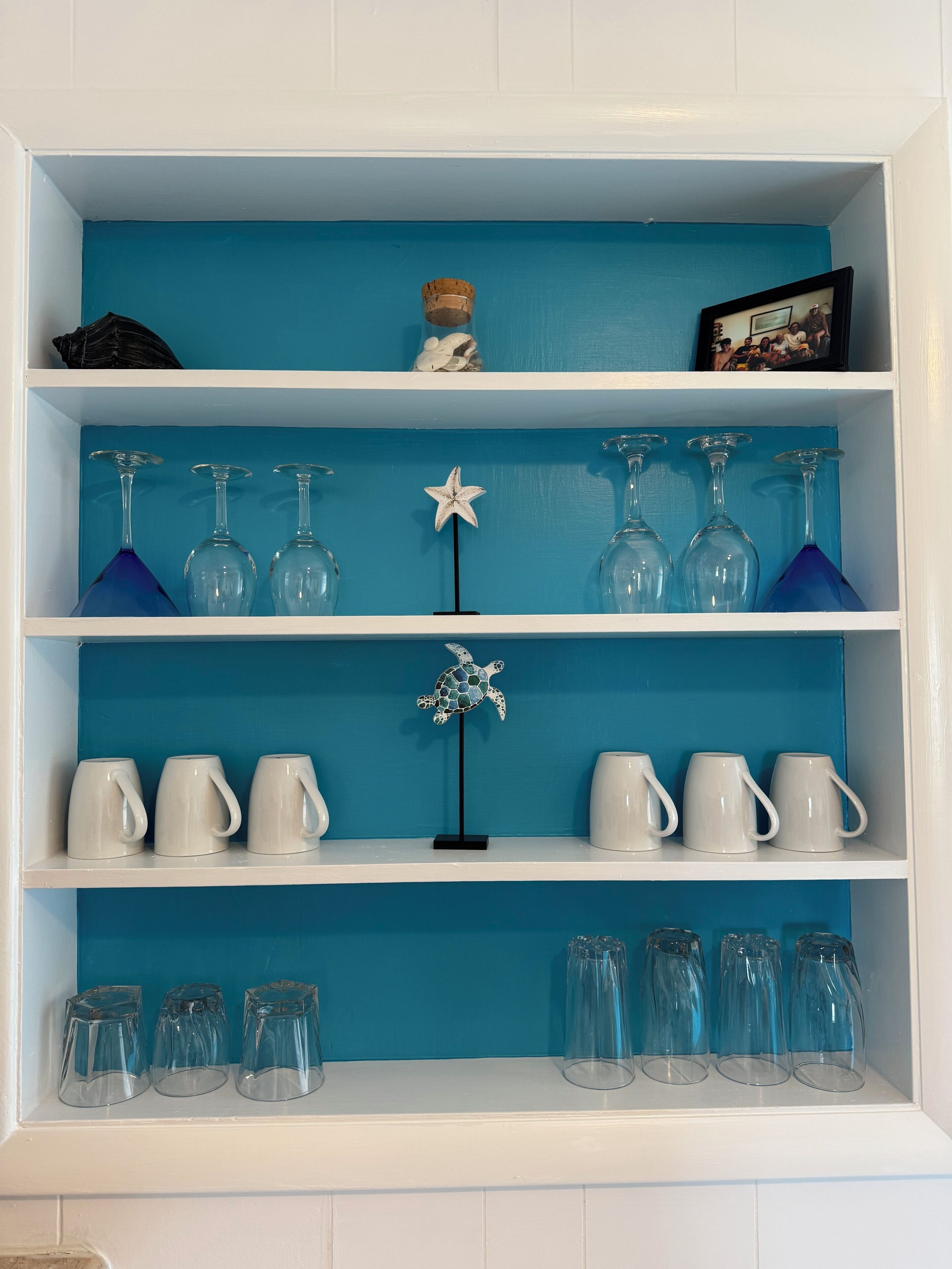 Une étagère peinte en bleu, ornée de verres à vin, de tasses blanches et d’une étoile de mer décorative.