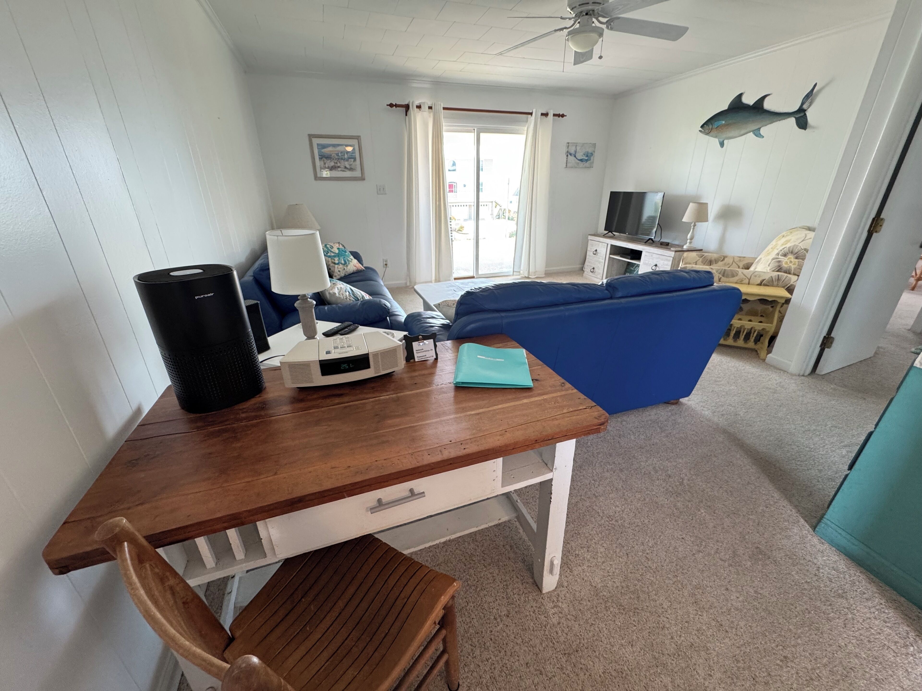 A living room with a wooden table, a blue sofa, a ceiling fan, and a fish decal on the wall.