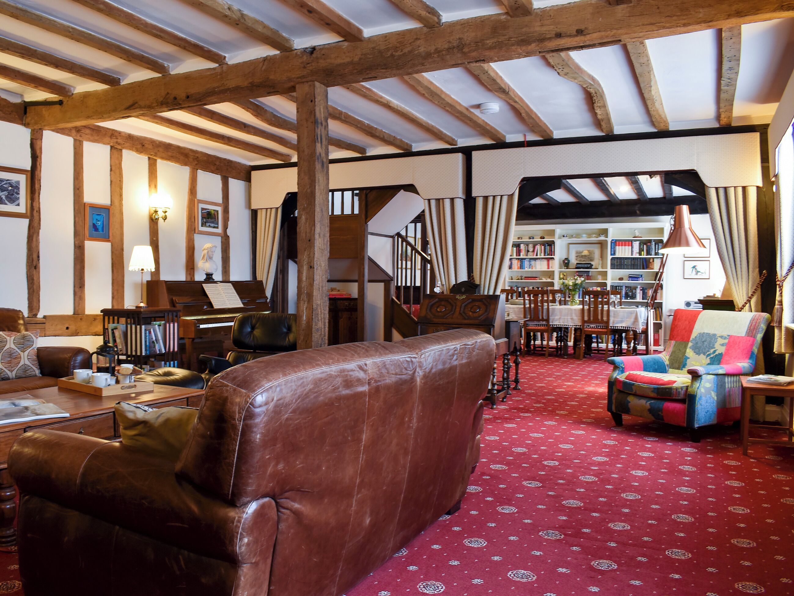 A cozy living room with exposed wooden beams, a piano, a bookshelf, and a red patterned carpet.