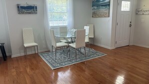 A dining area with a glass table and four chairs, a wooden floor, a decorative rug, and artwork on the walls. - Sunny Place Cottage,  Your Pet-Friendly Home away from Home! 🐾 (Colonial Beach)