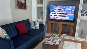 A living room with a blue sofa, a white fluffy rug, a wooden coffee table with a colorful cover, a television on a wooden stand, and a white fireplace mantel with a sign. - Sunny Place Cottage,  Your Pet-Friendly Home away from Home! 🐾 (Colonial Beach)