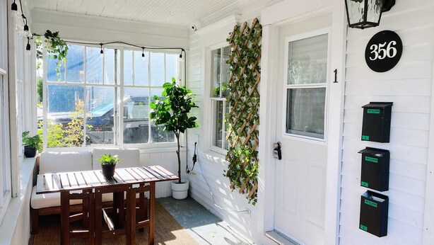 Varanda ensolarada com mesa e cadeiras de madeira, um vaso de planta e uma porta com o número 356.