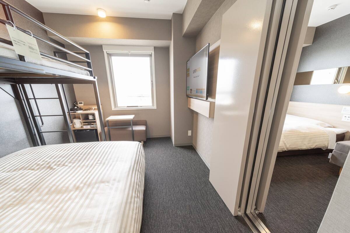 Een hotelkamer met een stapelbed, een bureau met een stoel, een klein raam en een bed met een gestreept dekbed.
