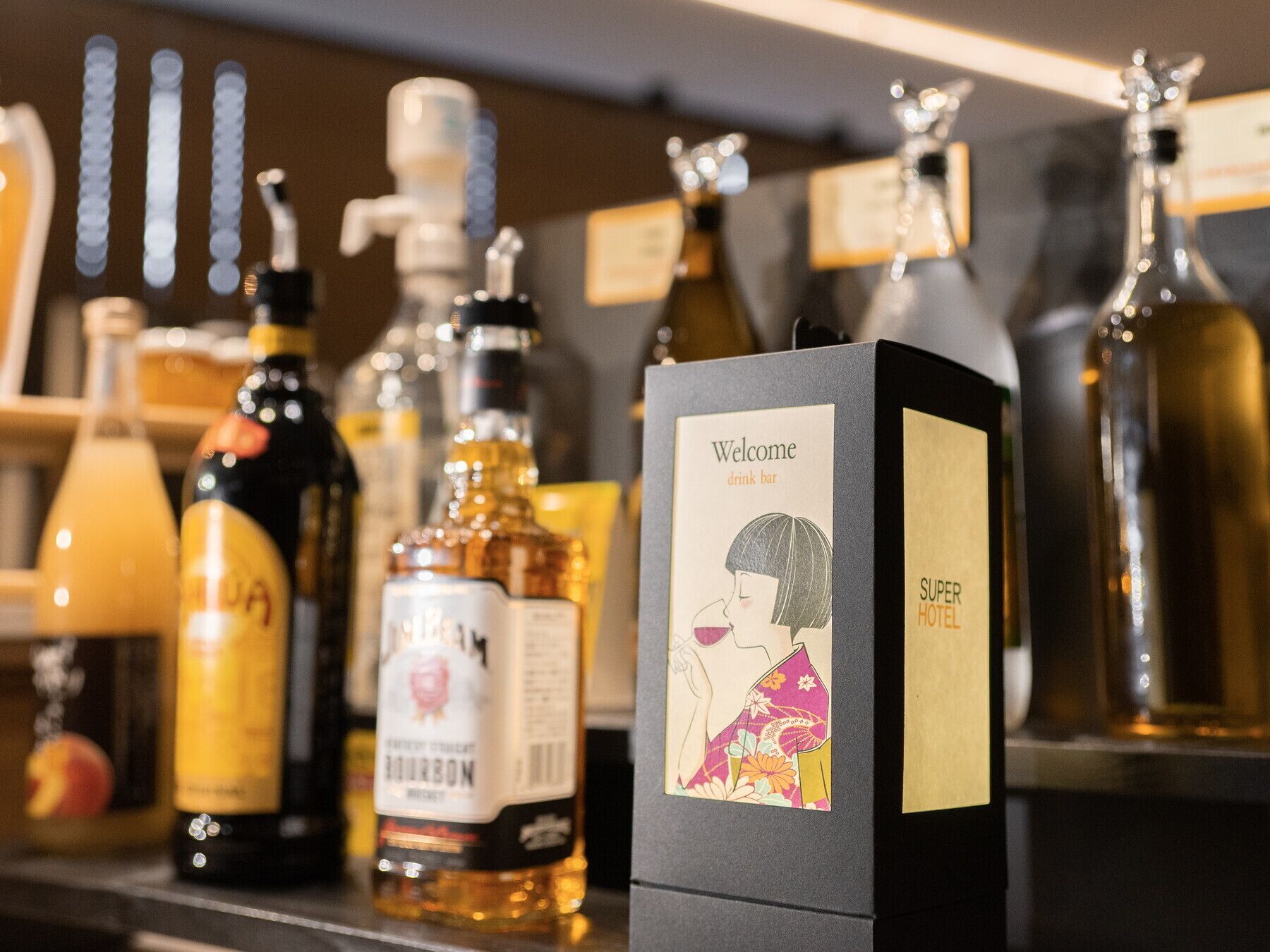 Varias botellas de licor en un estante con un letrero que indica una barra de bienvenida y la etiqueta 'Super Hotel'.
