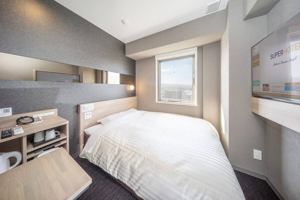 Hotelkamer met een bed, een bureau met telefoon en waterkoker, een spiegel en een raam met uitzicht op de stad.