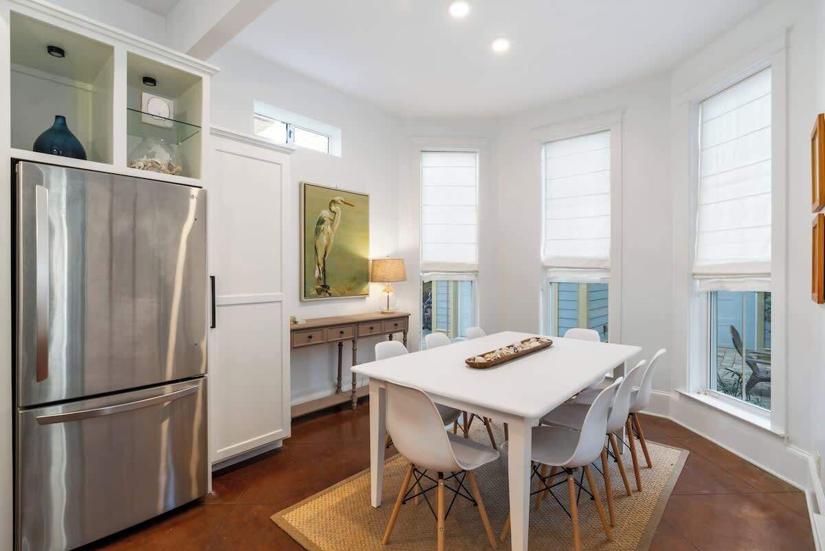 A modern kitchen with a stainless steel refrigerator, white cabinetry, a dining table with chairs, and large windows.