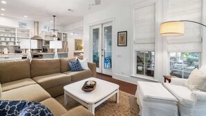 Amplia sala de estar con sofá color beige, una mesa de centro blanca y un sillón blanco. Se observa una zona de cocina con armarios blancos y un ventilador de techo.