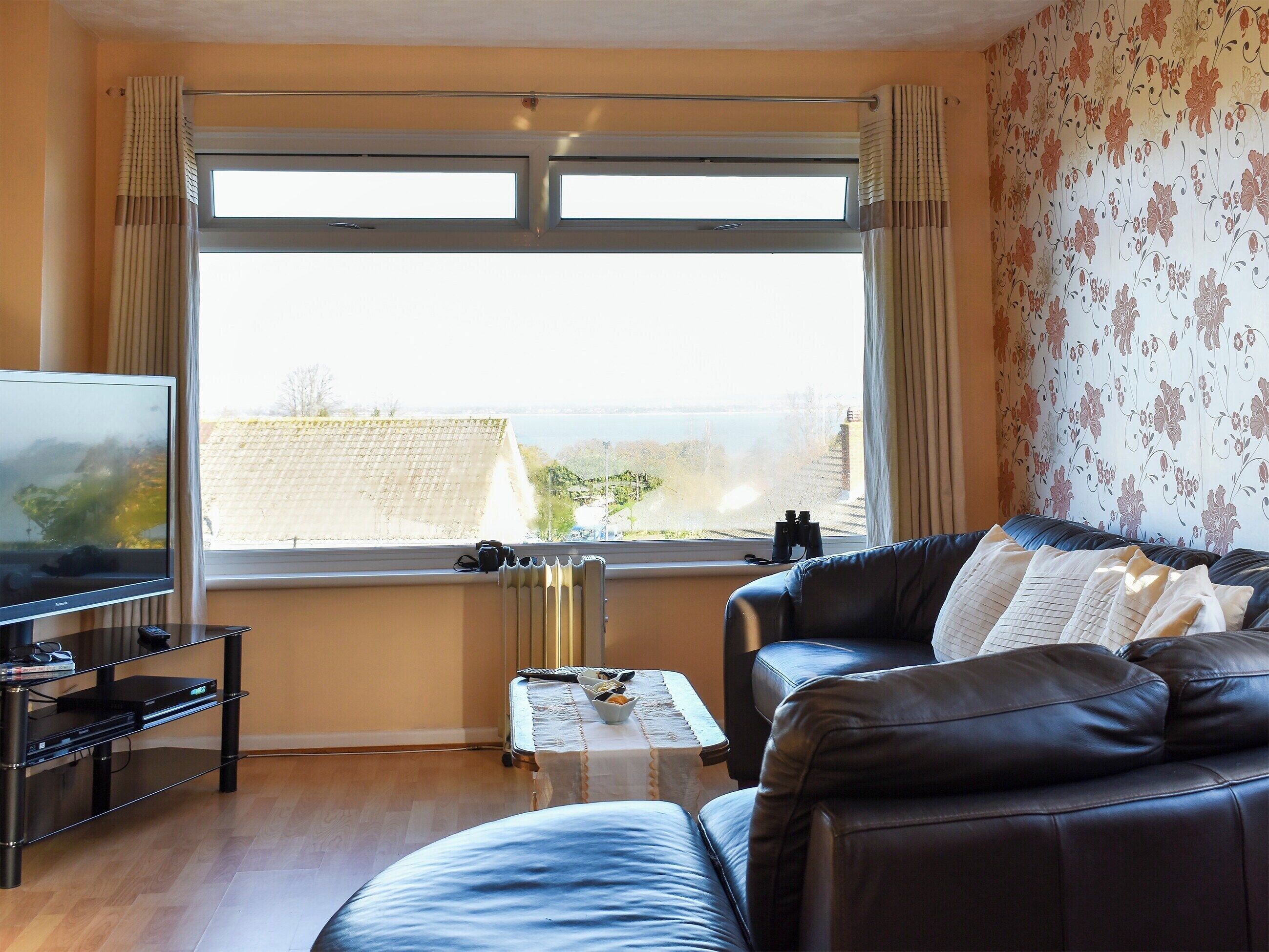 Ein Wohnzimmer mit Fernseher, einem Ledersofa, einem Couchtisch und Blick auf ein Gewässer durch große Fenster.