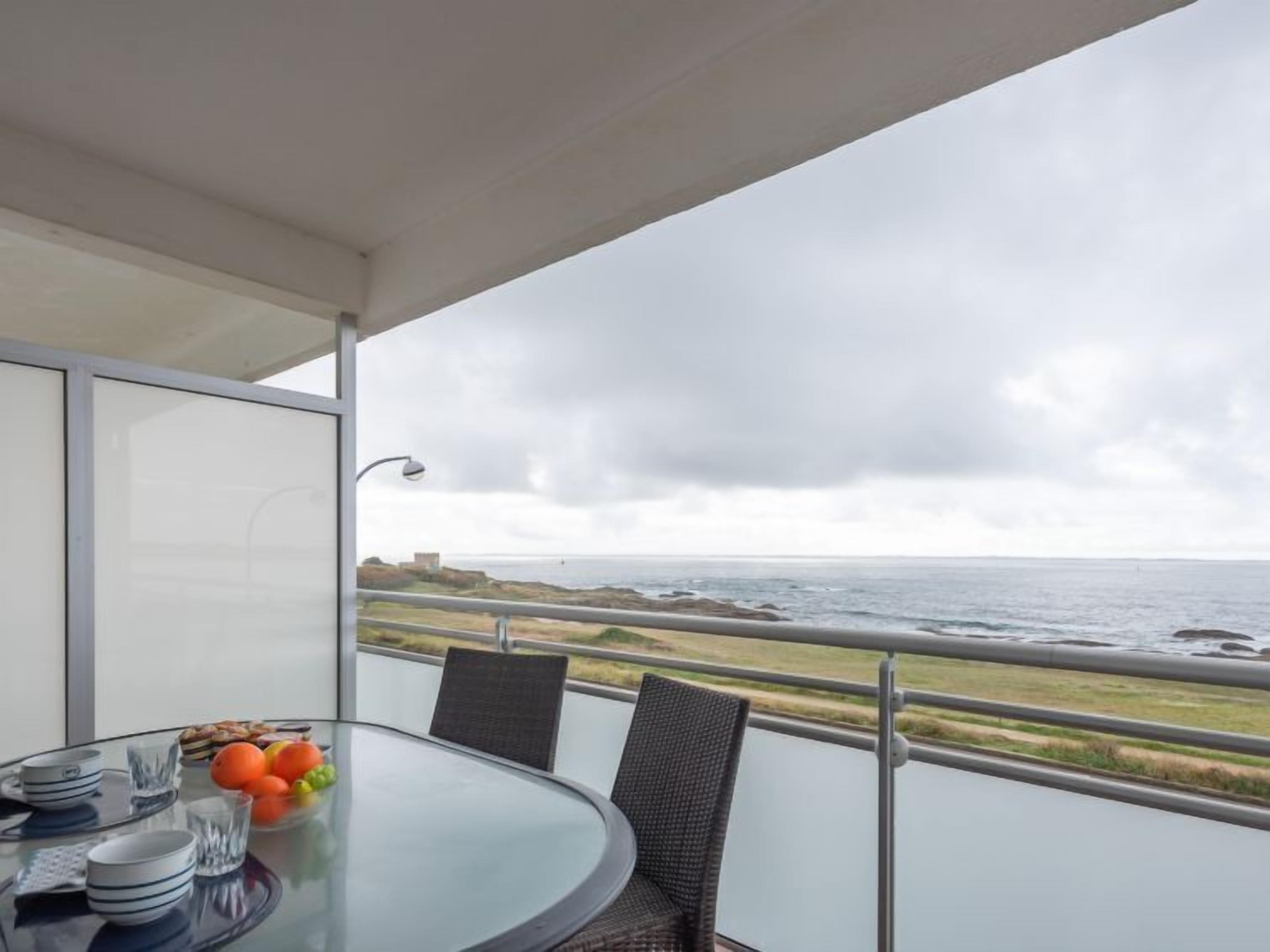 Varanda com mesa de vidro posta para refeições, com vista para a costa, mar e área ajardinada.