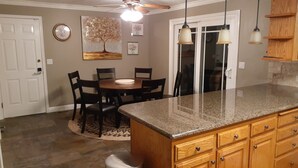 A kitchen with granite countertops, wooden cabinets, a dining table with chairs, a ceiling fan, and a wall clock.