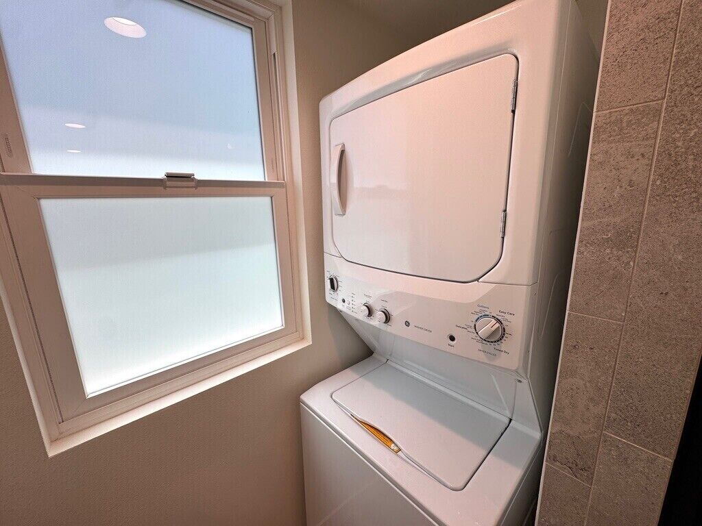 Un lavadero pequeño con lavadora y secadora apiladas, una ventana y pared de azulejos.
