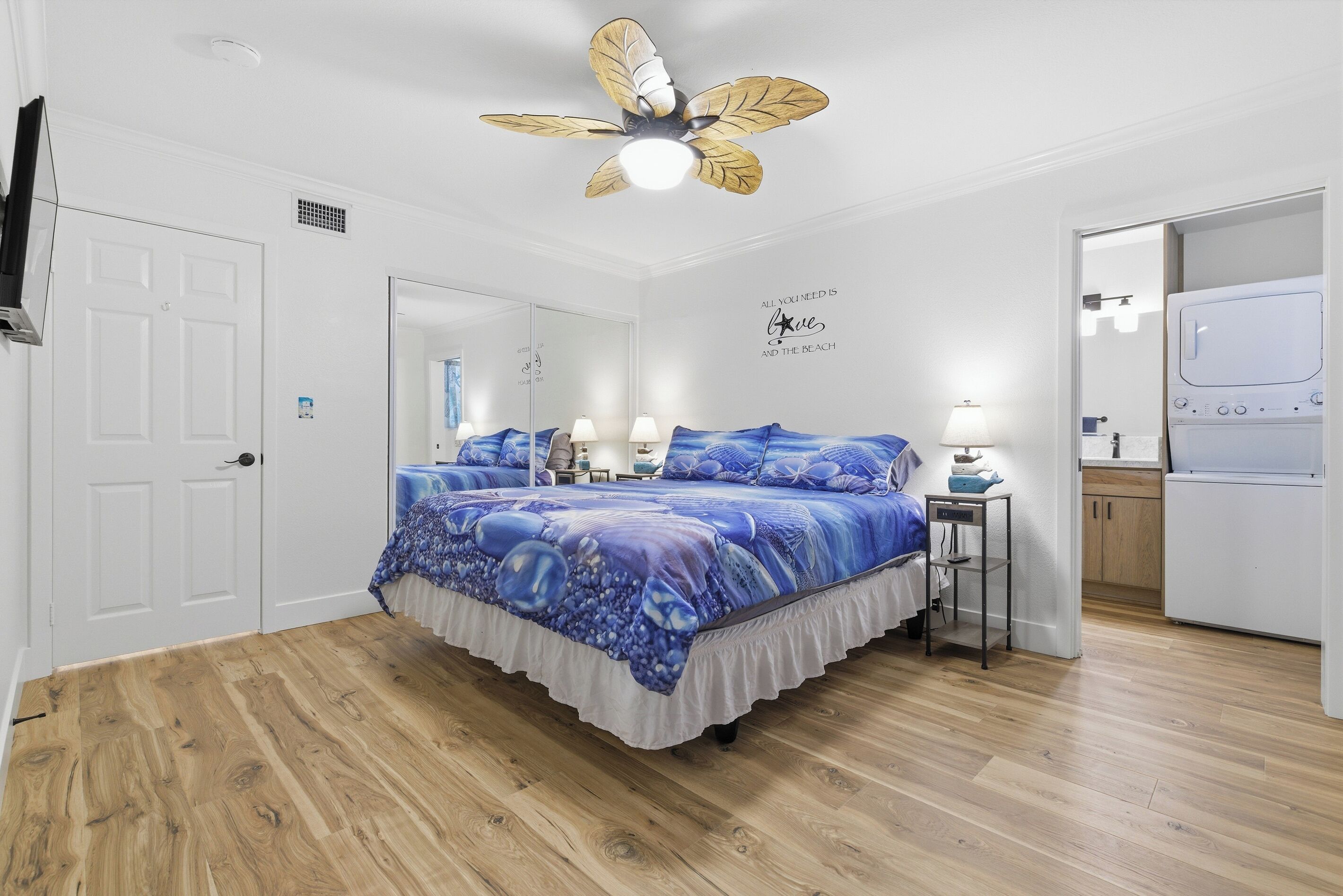 Un dormitorio con cama, ventilador de techo, espejo, pared con cita motivacional y zona de lavandería.