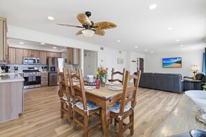 A modern kitchen with wooden cabinets, a dining area with a wooden table and chairs, a ceiling fan, and a living room with a sofa and a TV.