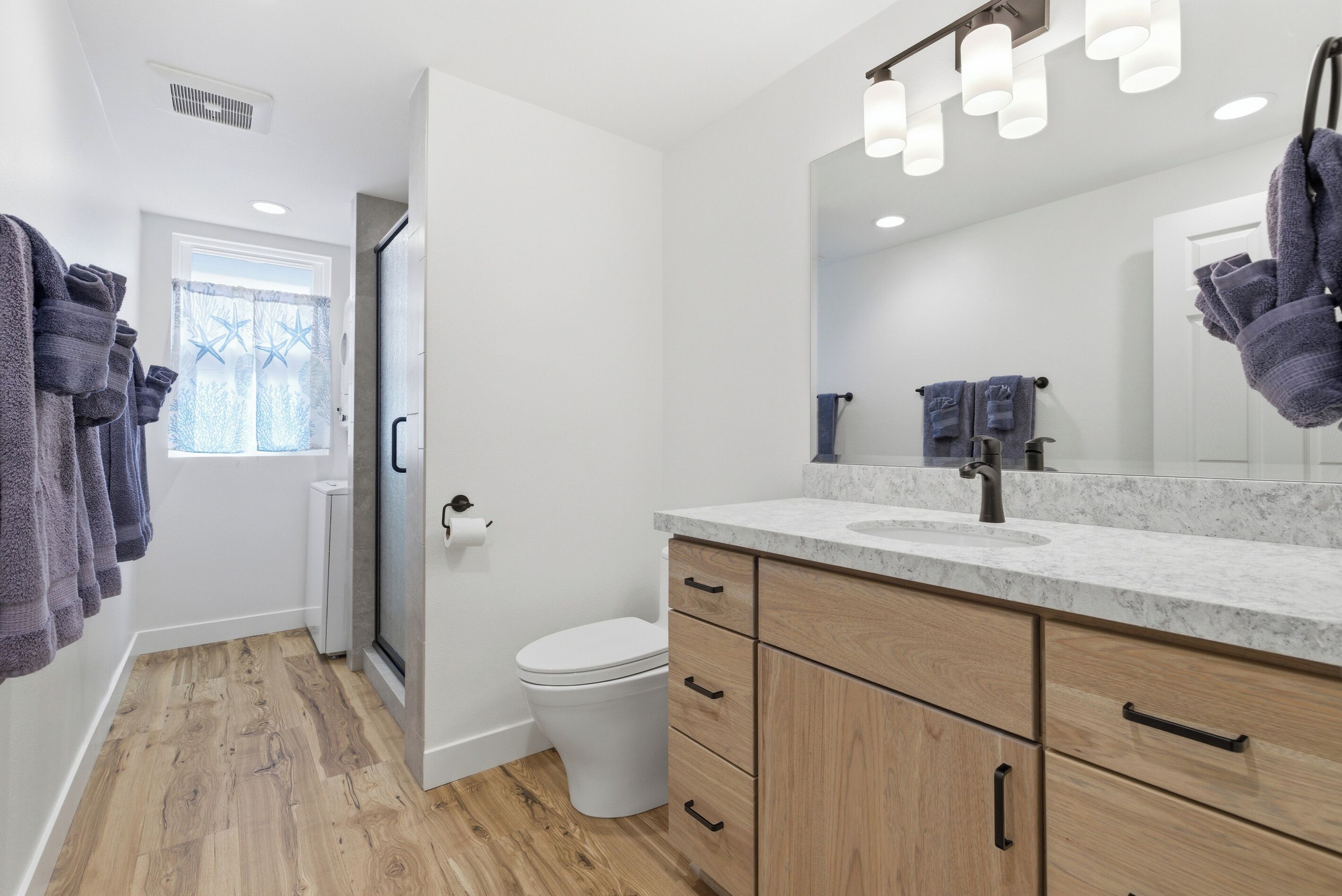 A modern bathroom with a wooden floor, a white toilet, a marble sink, and a large mirror with lights.