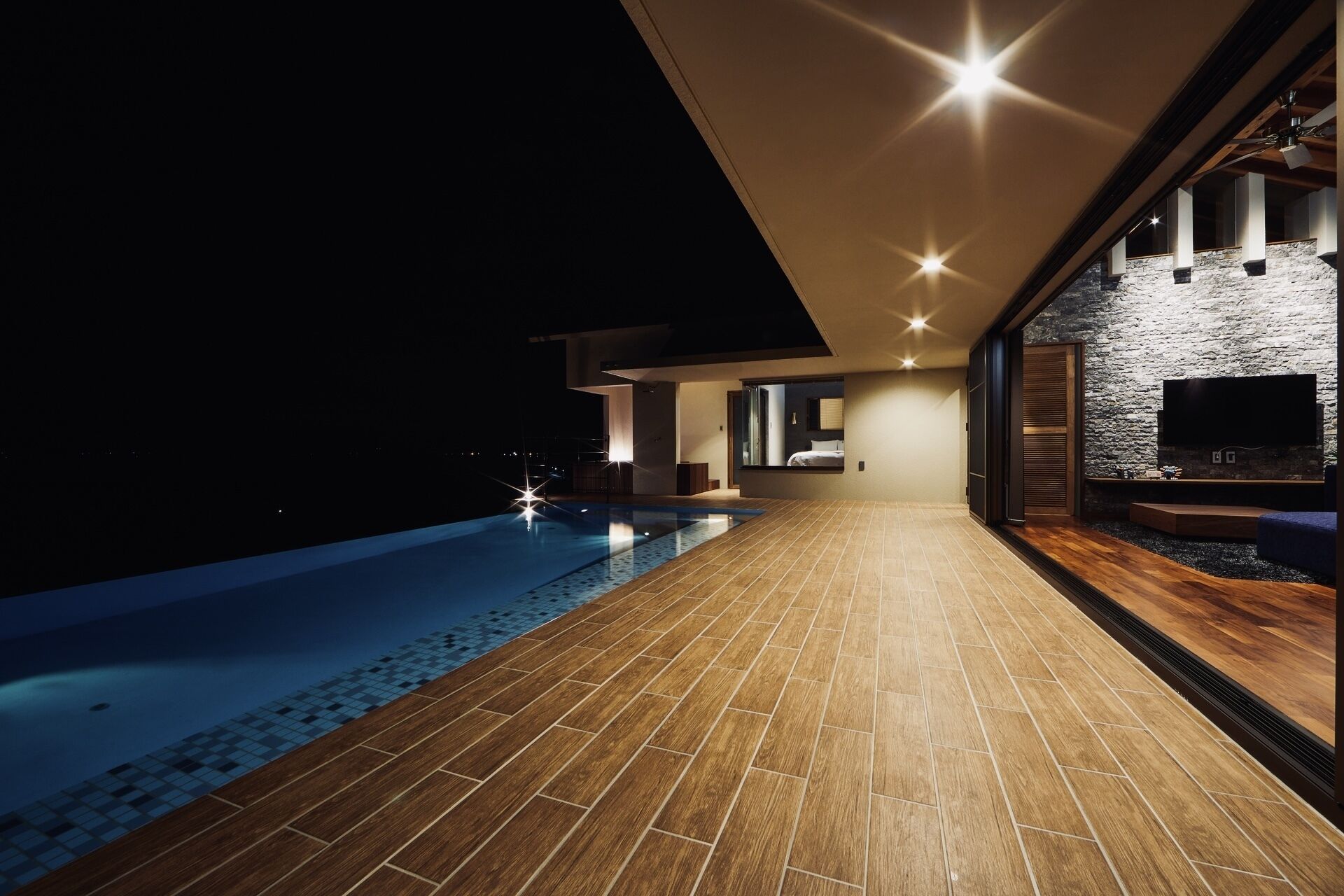 A modern pool area with a wooden deck leading to a stone wall with a TV.