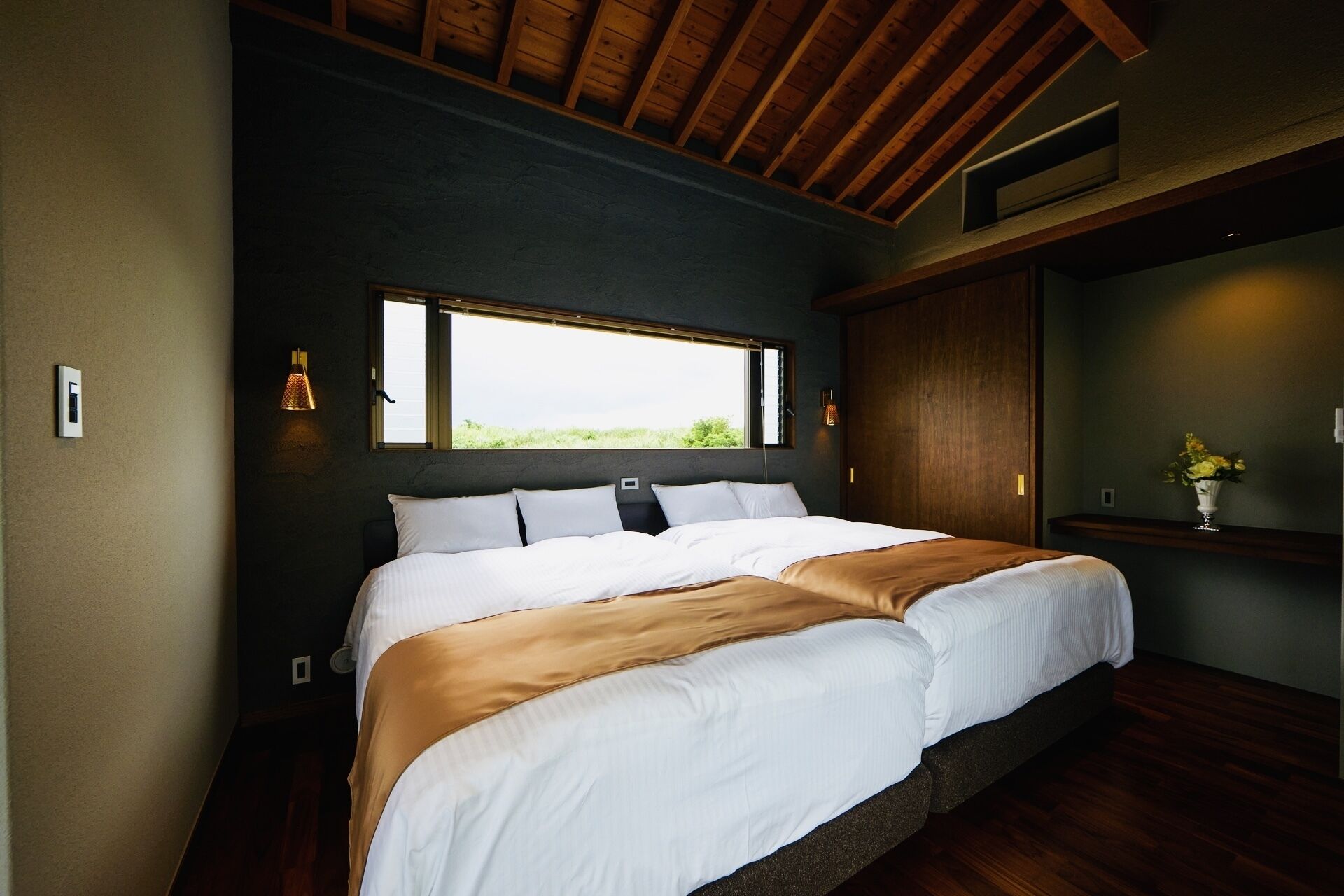 A bedroom with a large bed, a window, a wooden ceiling, and a nightstand with a vase.