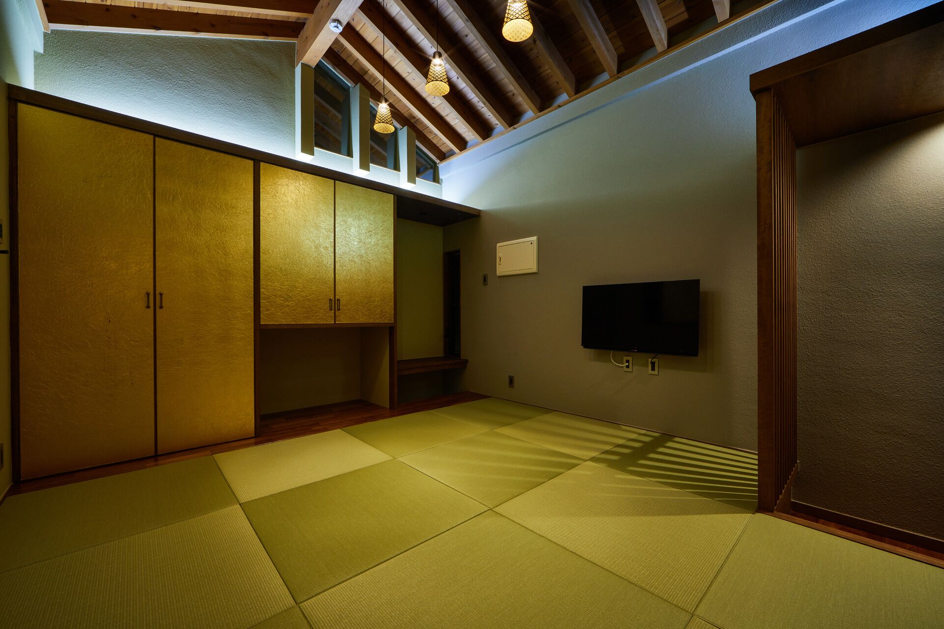 A room with tatami mats, wooden cabinets, a television, and a ceiling with exposed beams and hanging lights.